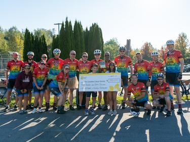Group photo of Tour de Valley participants posing for photo with two students holding a giant cheq.